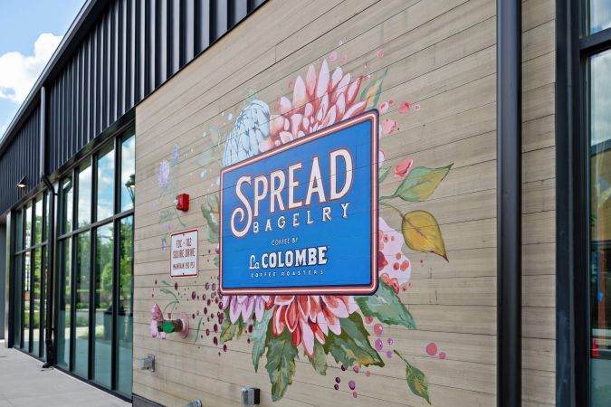 A colorful "Spread Bagelry" mural with flowers on an exterior wall next to large glass windows.
