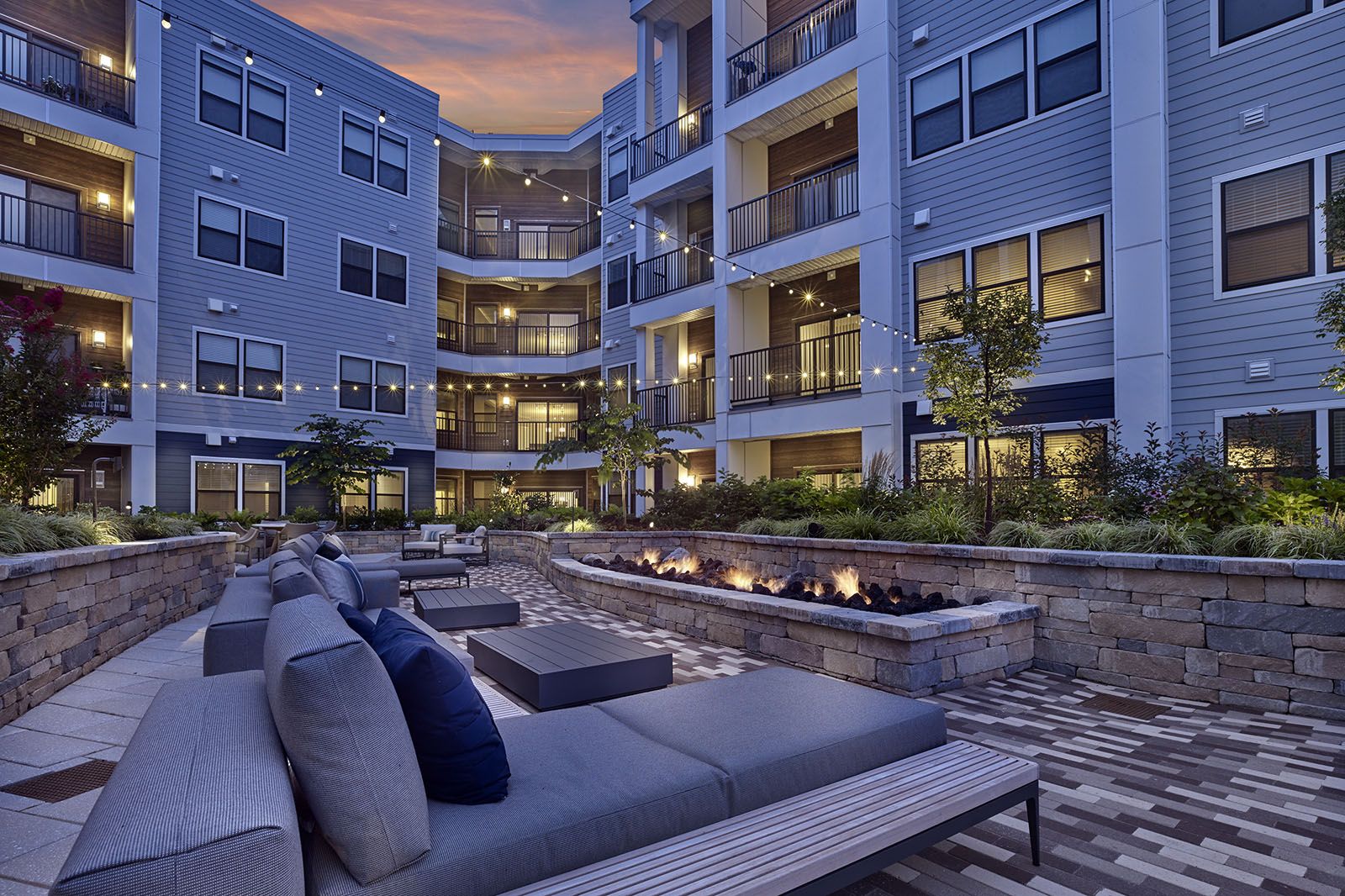 Modern apartment courtyard at dusk with lounge seating, fire pit, string lights, and landscaped garden.