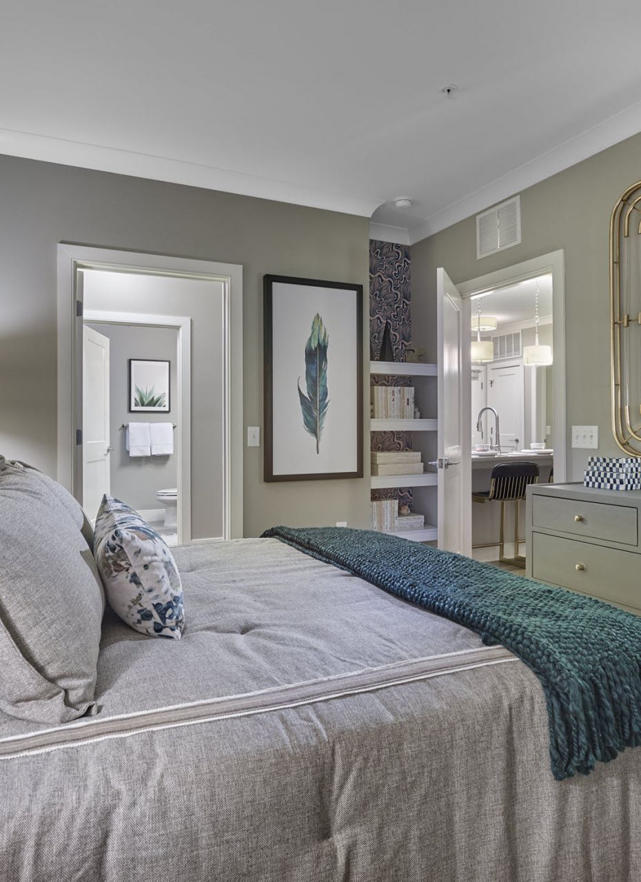 Modern bedroom with gray bedding, green and blue pillows, dresser, mirror, and open doorways to other rooms.