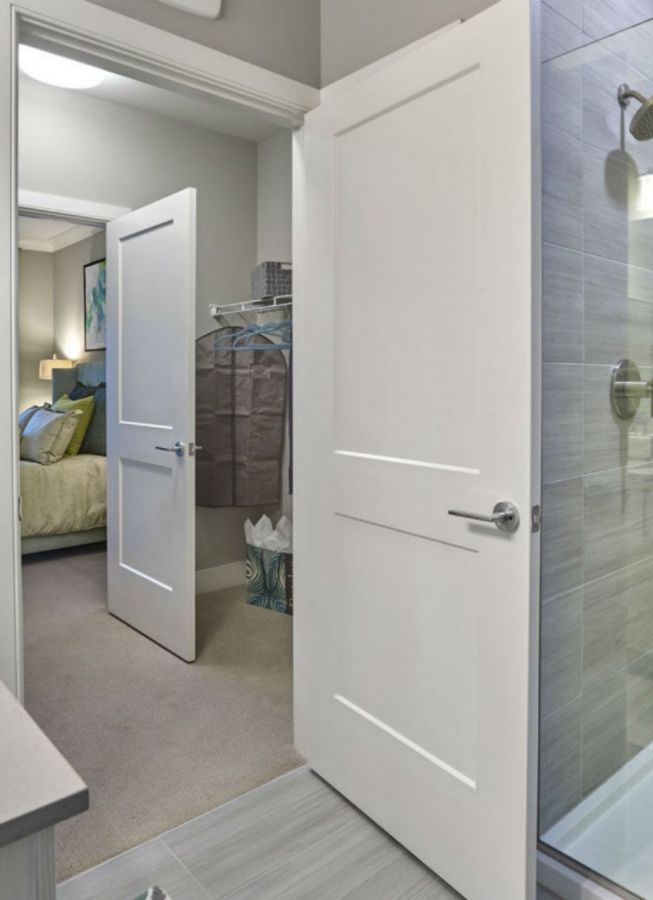 Modern bathroom with a glass shower, gray countertops, and a view into a bedroom with a bed and closet.