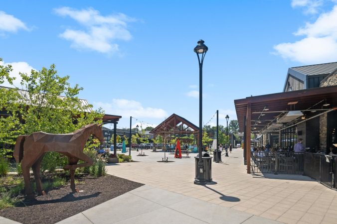 Outdoor shopping area with a metal horse sculpture, patio dining, and people walking under a sunny, blue sky.
