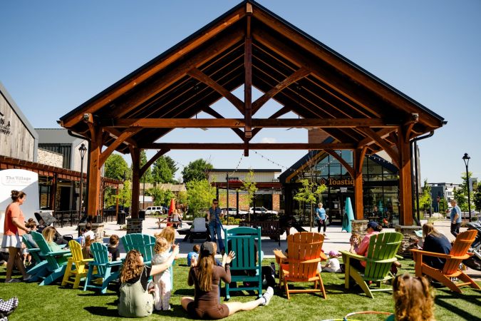 People sit in colorful chairs on grass under a wooden pavilion at an outdoor community event on a sunny day.
