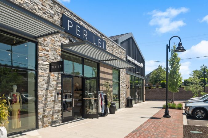 Boutique storefronts with stone exteriors and large windows on a sunny day, clothes displayed outside.