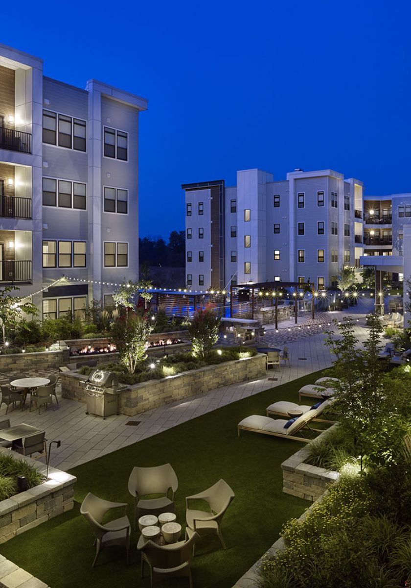 Modern apartment complex courtyard at dusk, with seating areas, greenery, and string lights illuminating the scene.