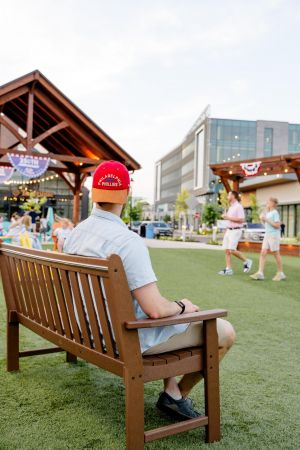 A man in a red hat sits on a bench in a lively outdoor plaza with people walking and socializing.