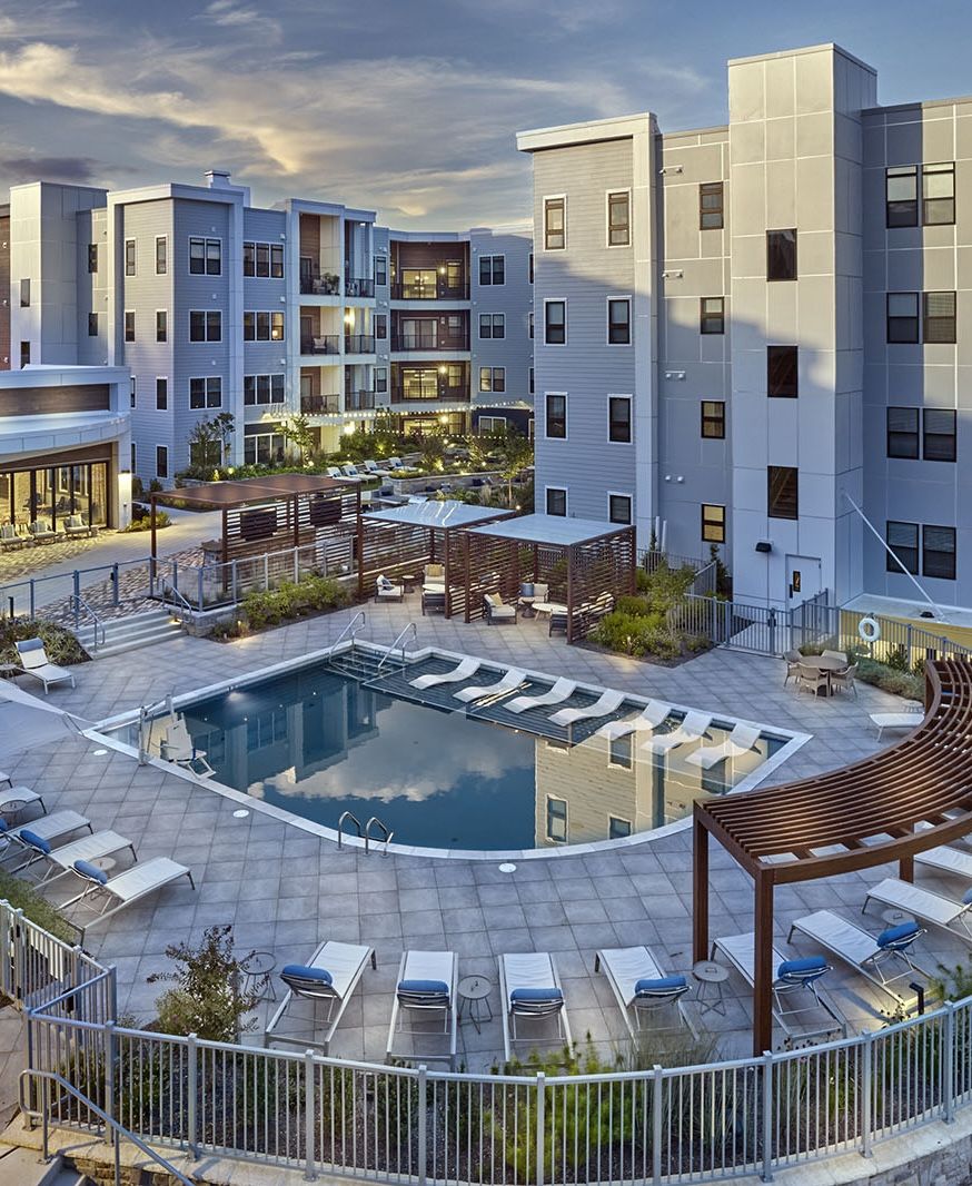 Modern apartment complex with a central outdoor pool, lounge chairs, and shaded seating areas at sunset.