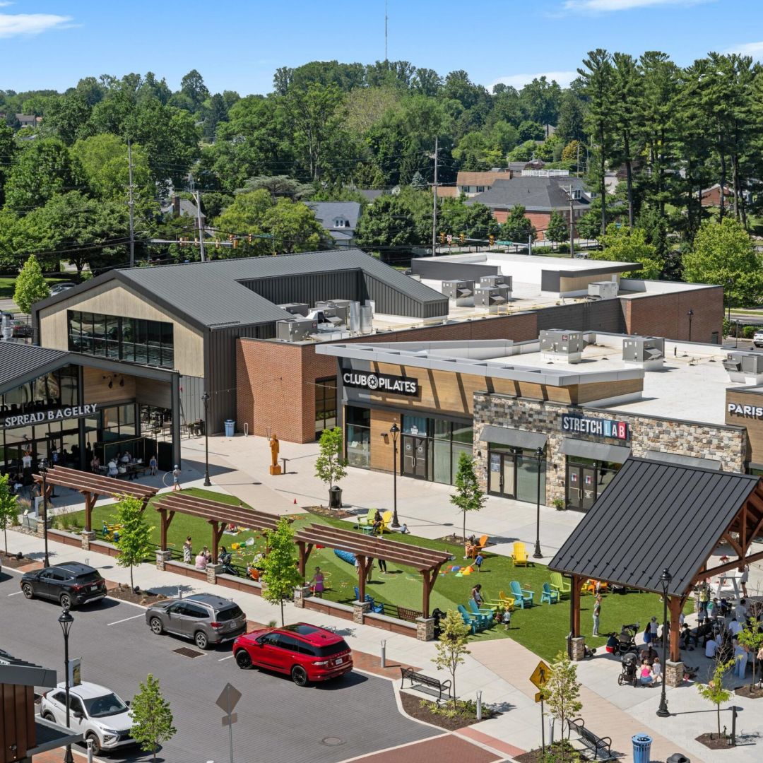 A modern shopping plaza with shops, green spaces, and people gathered under a pavilion on a sunny day.