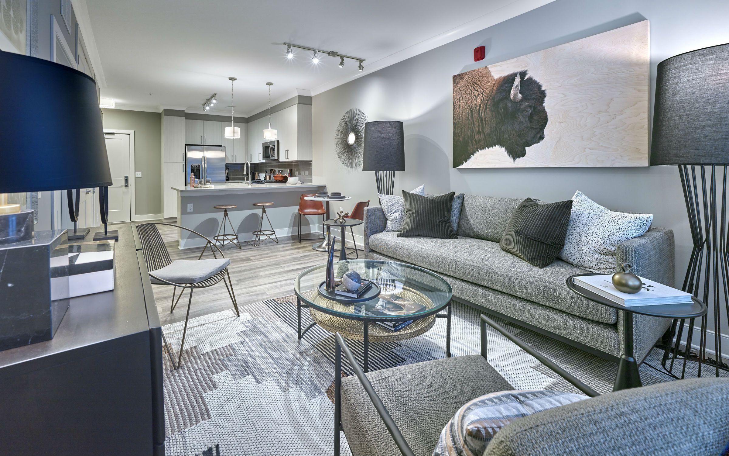 Modern living room with gray furniture, glass coffee table, and open kitchen in the background.