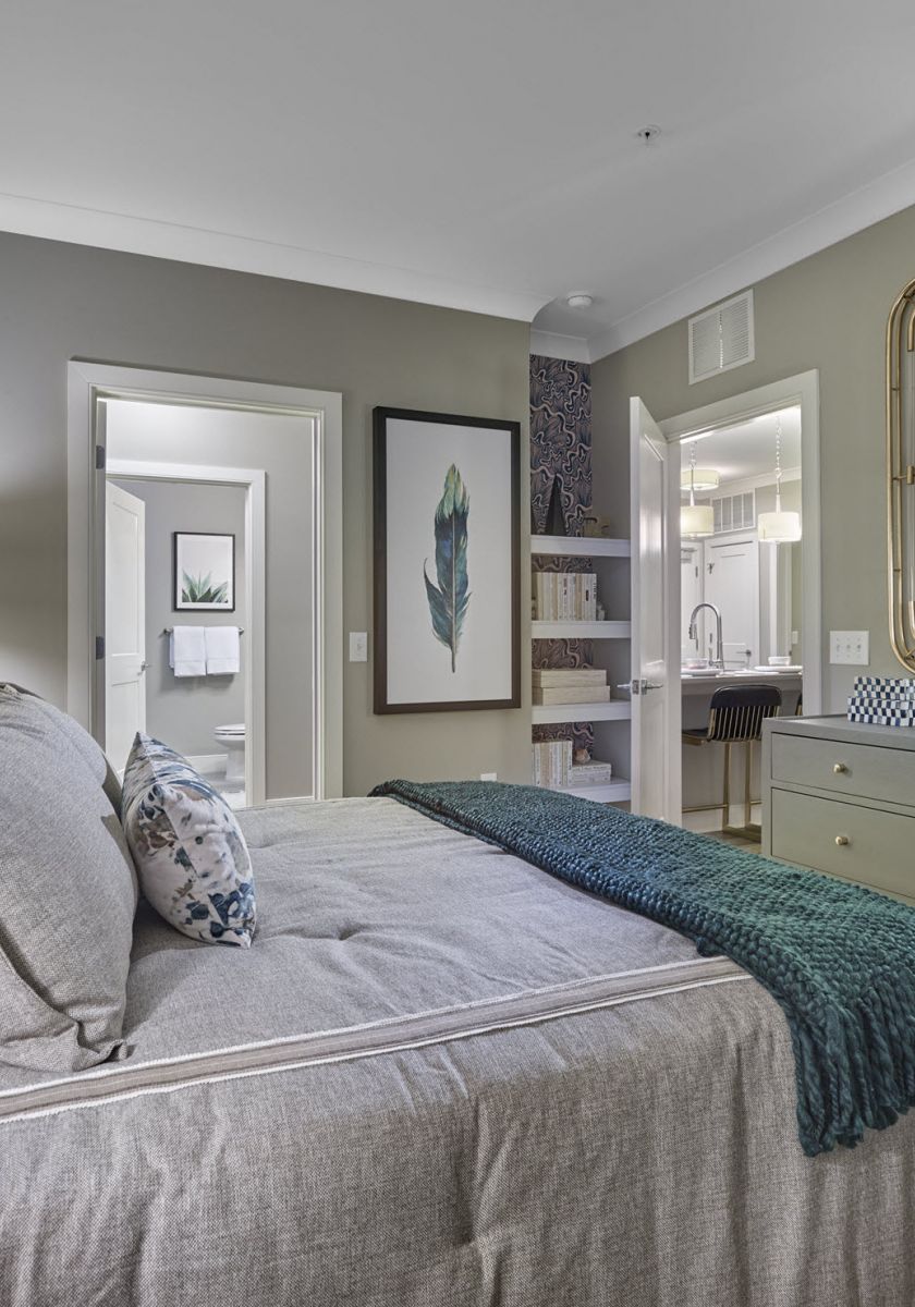 Modern bedroom with gray bedding, green and blue pillows, dresser, mirror, and open doorways to other rooms.
