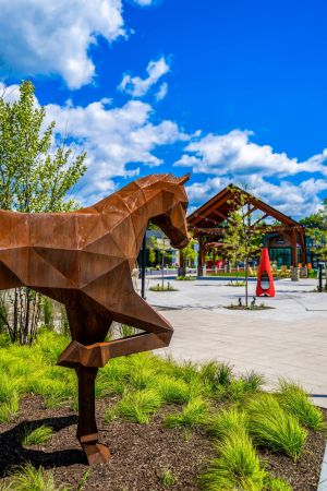A geometric metal horse sculpture stands among greenery near a pavilion under a bright blue sky with clouds.