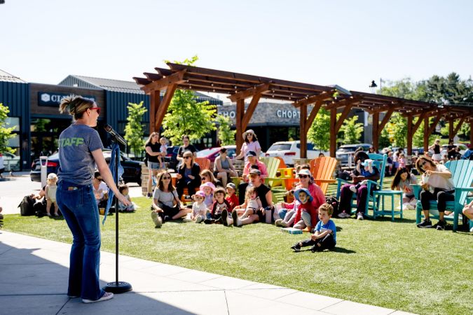 A staff member speaks to a group of children and adults sitting outdoors on grass and colorful chairs.