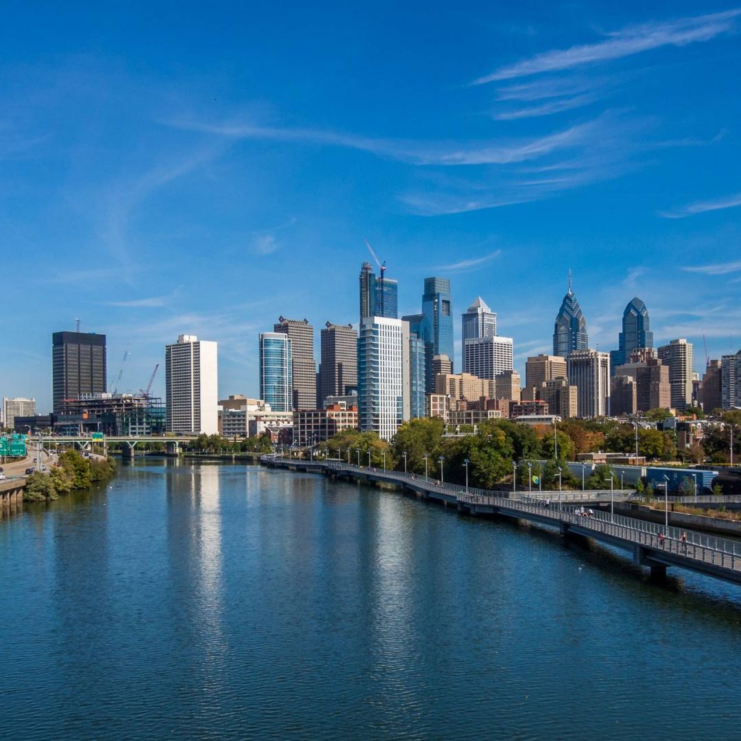 Philadelphia skyline with modern skyscrapers reflected in the Schuylkill River under a clear blue sky.