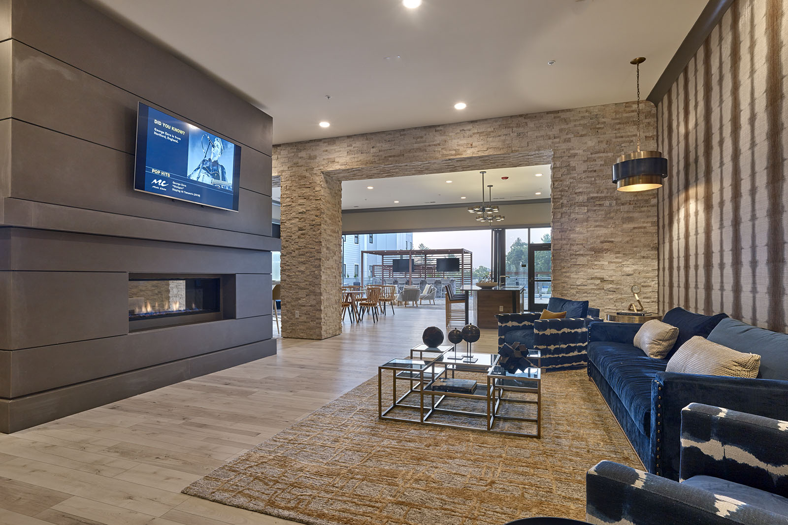 Modern lounge with blue seating, glass tables, a fireplace, wall-mounted TV, and dining area in the background.