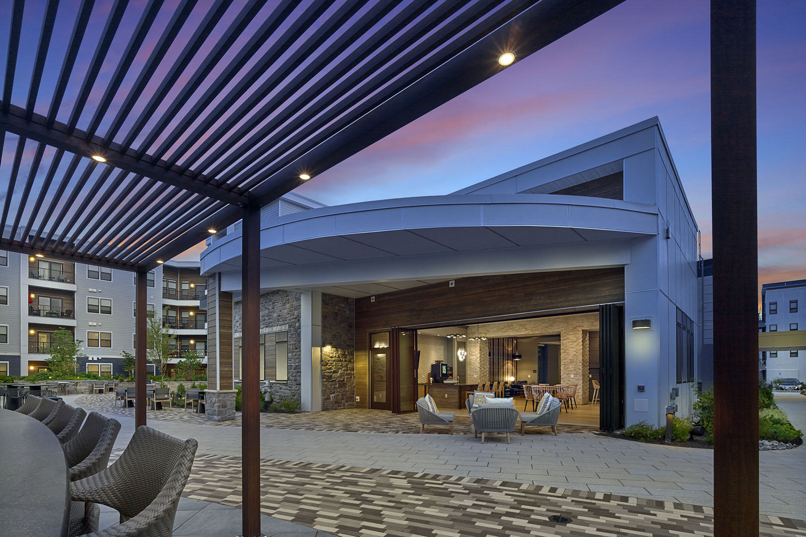 Modern outdoor patio with seating, pergola, and large glass doors opening to an indoor lounge at sunset.