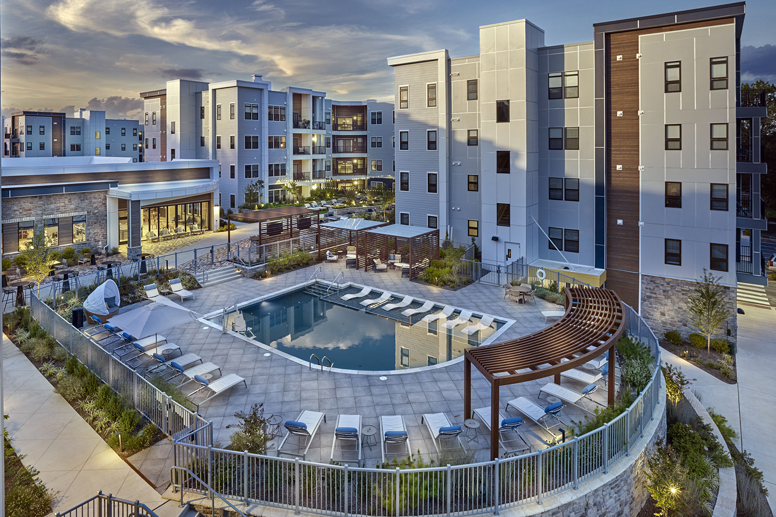 Modern apartment complex with a central outdoor pool, lounge chairs, and pergolas at sunset.