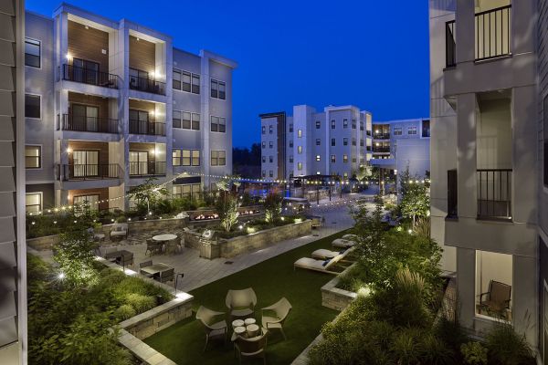 Modern apartment complex courtyard at dusk, with seating areas, greenery, and string lights illuminating the scene.