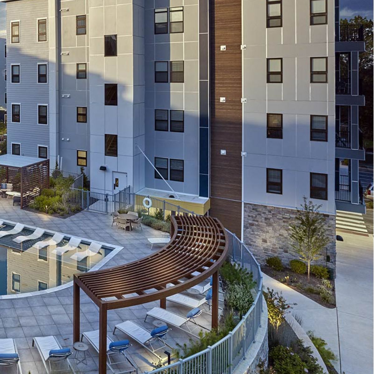 Modern apartment building with a pool, lounge chairs, and a curved wooden pergola in the courtyard.