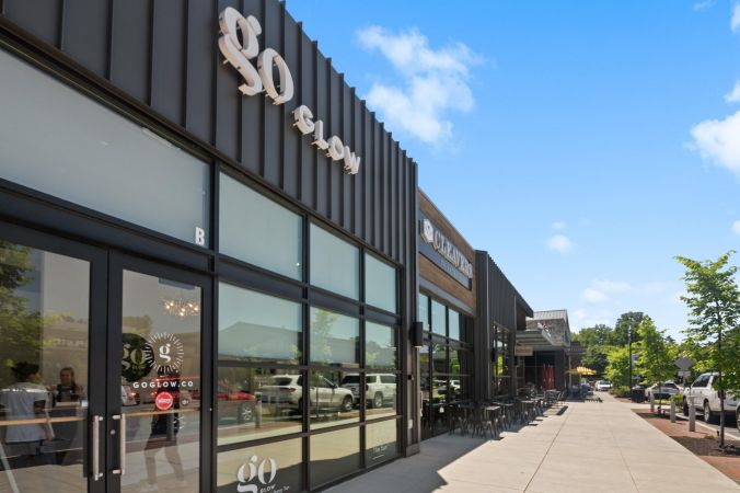 Street view of modern storefronts with large glass windows and outdoor seating on a sunny day.