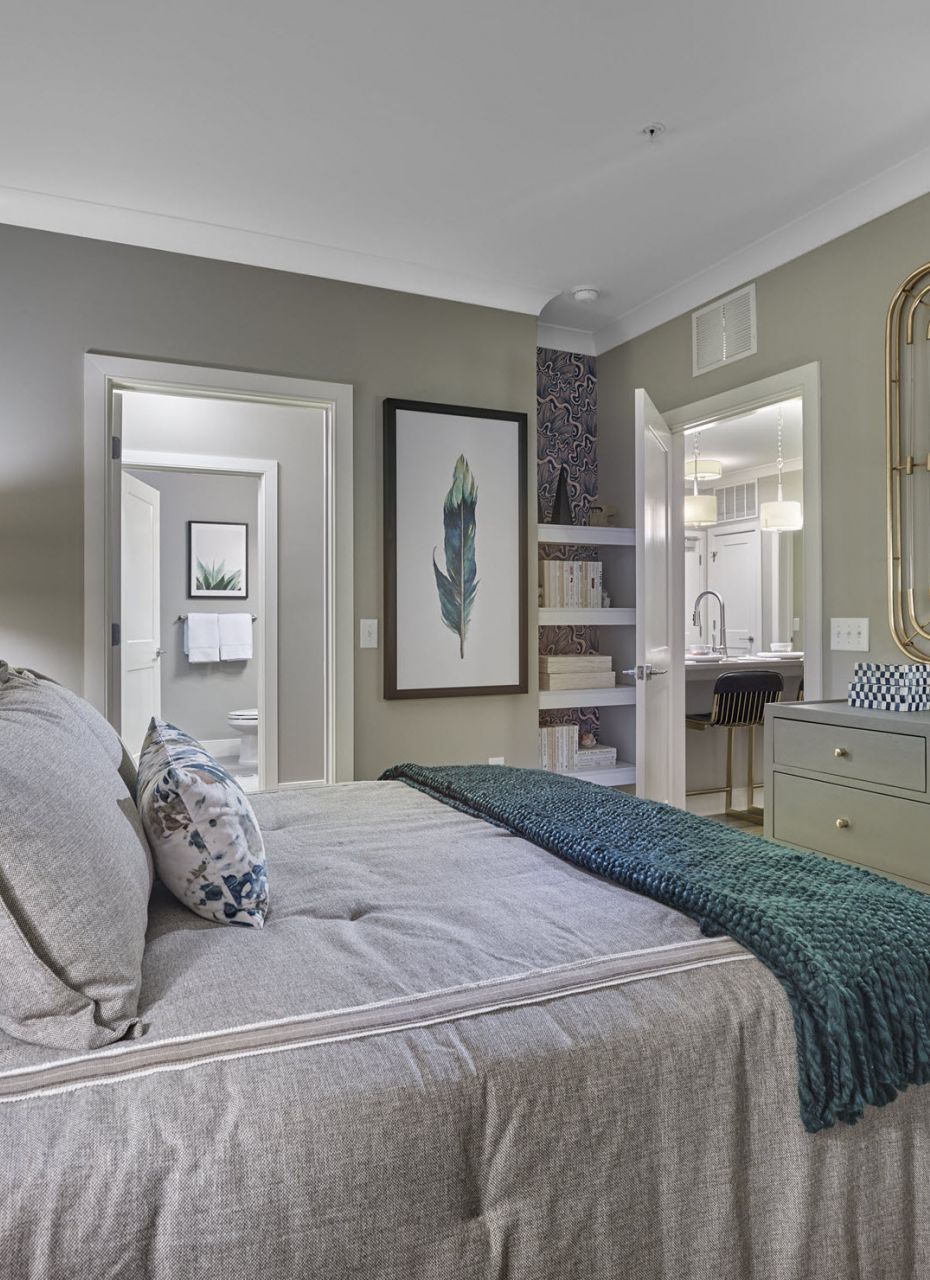 Modern bedroom with gray bedding, green and blue pillows, dresser, mirror, and open doorways to other rooms.
