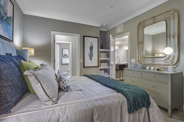 Modern bedroom with gray bedding, green and blue pillows, dresser, mirror, and open doorways to other rooms.