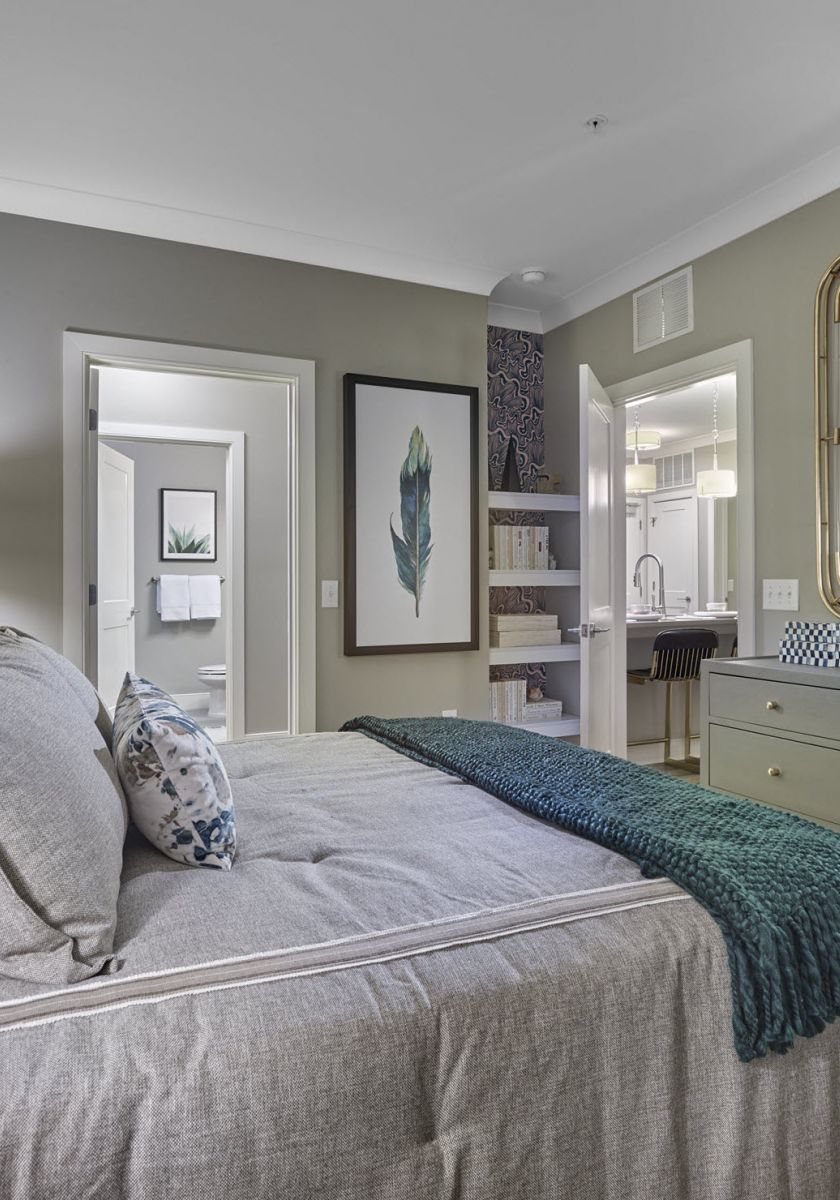 Modern bedroom with gray bedding, green and blue pillows, dresser, mirror, and open doorways to other rooms.