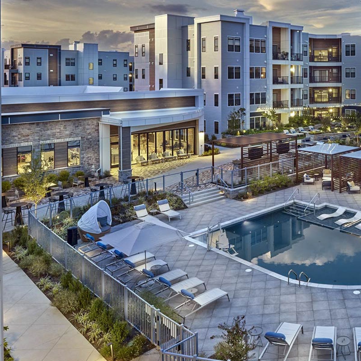 Modern apartment complex with outdoor pool area, lounge chairs, and landscaped surroundings at sunset.